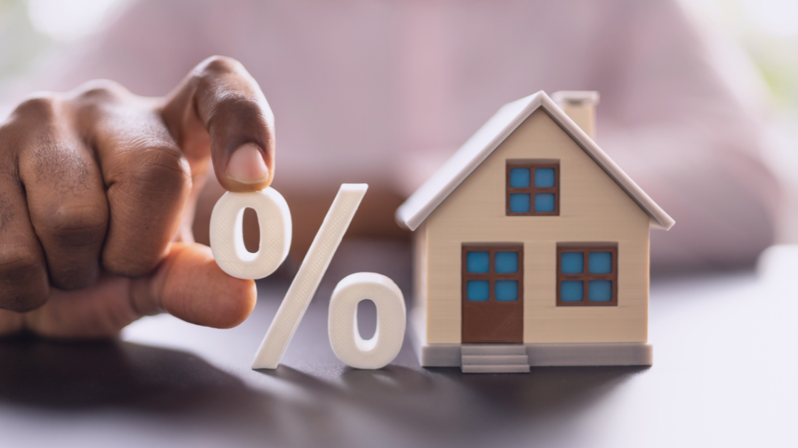 Should I Buy a House in My Company, in a Trust, or in My Personal Name? Close-up of a hand holding a percentage sign next to a small model house, symbolizing homeownership and mortgage decisions.