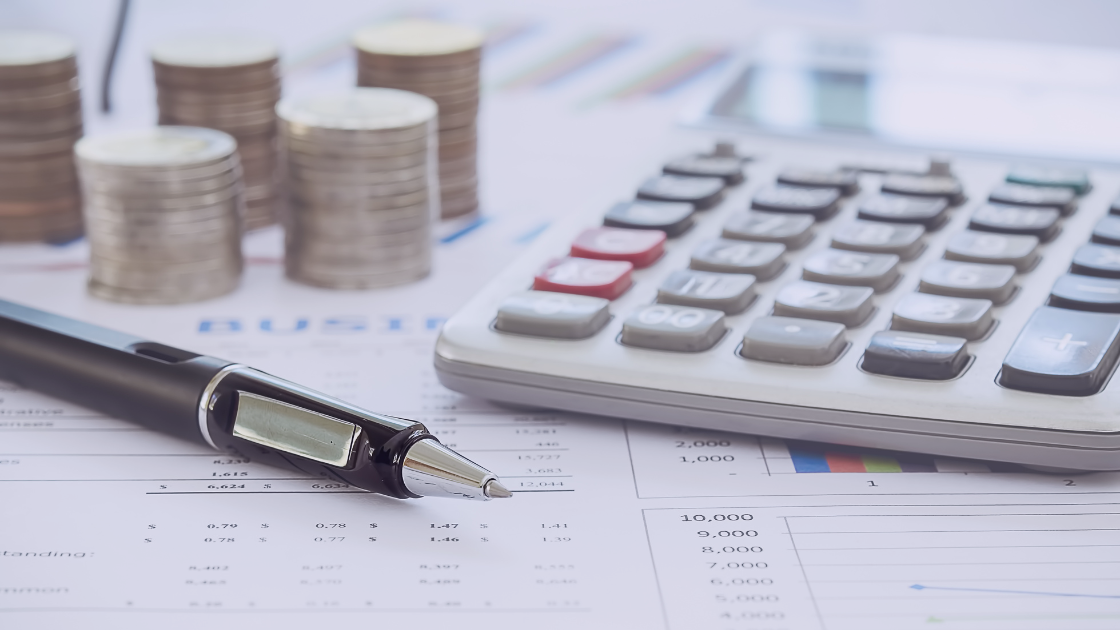 A close-up of a black pen, calculator, and stacked coins on financial documents with charts and tables, representing accounting, tax, and director remuneration planning.